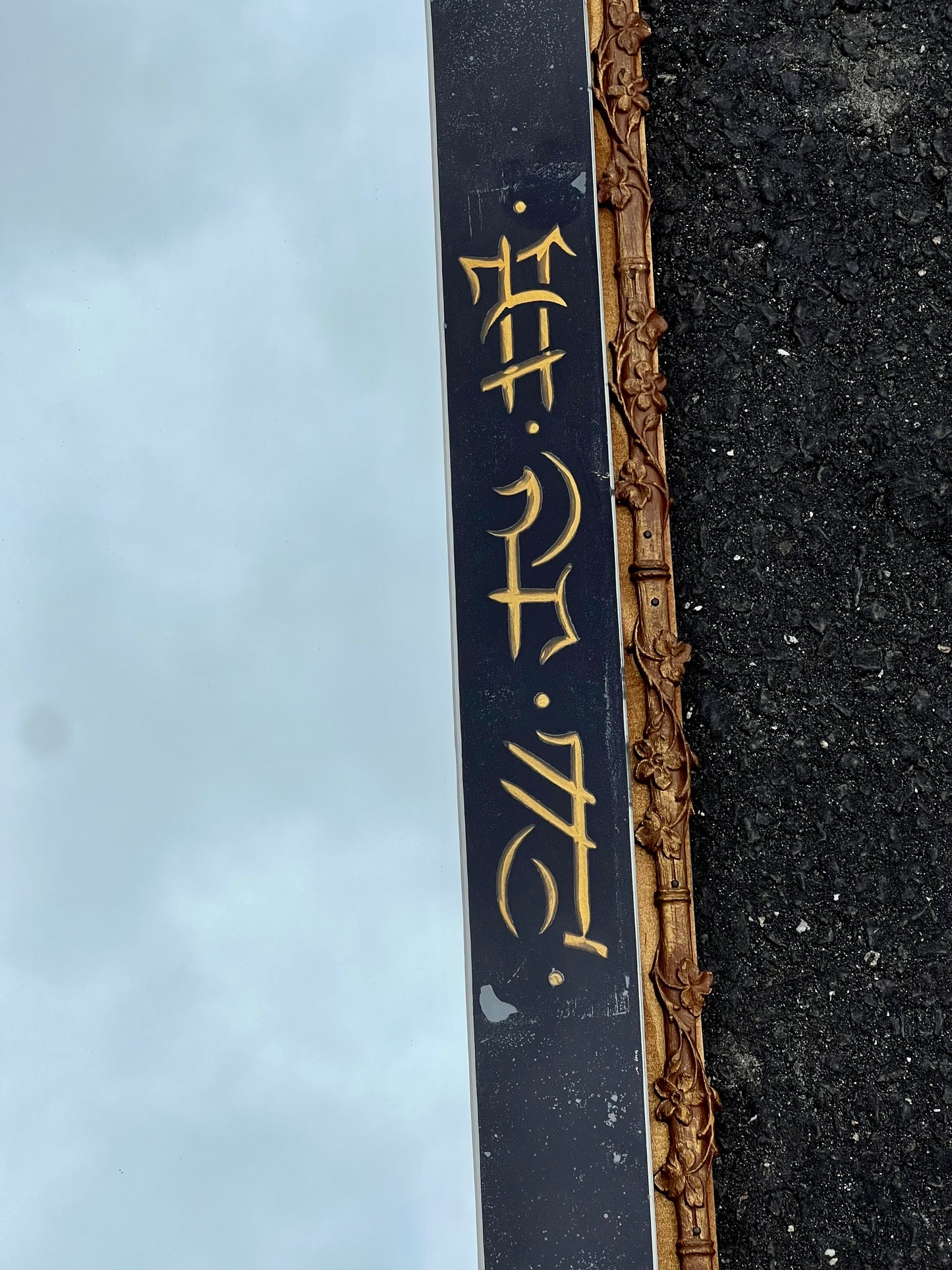 A close-up of the Pearl City Antiques Vintage Japanned Gilt Wood Wall Mirror, featuring a black frame, gold floral accents, and vertical script, lies on black asphalt reflecting a cloudy sky and elegant gilt detailing.