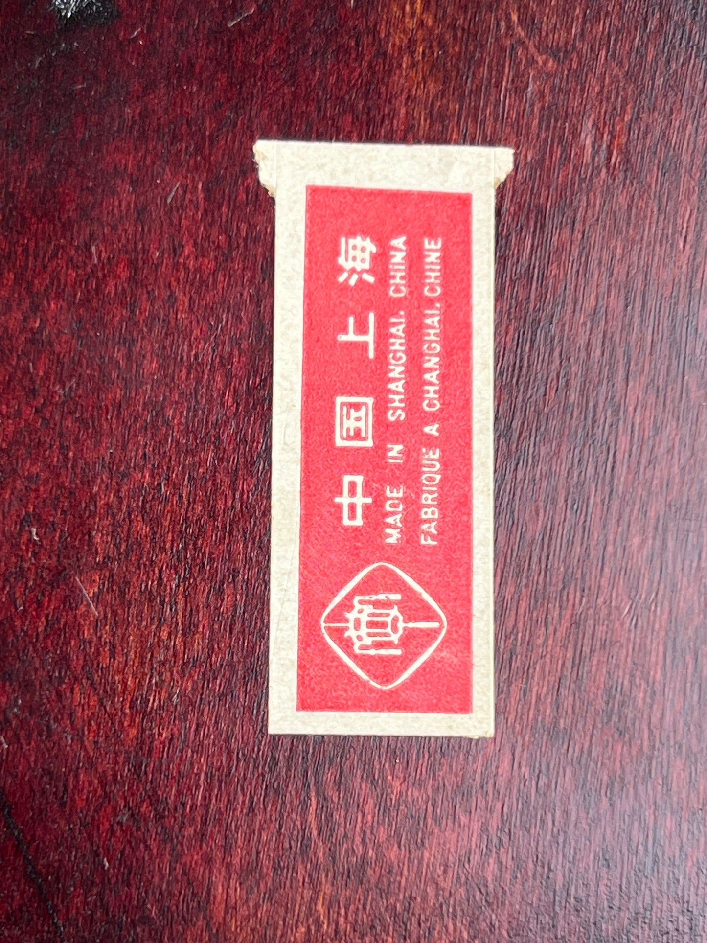 A rectangular red and beige label with Chinese characters and "MADE IN SHANGHAI, CHINA / FABRIQUÉ À SHANGHAI, CHINE" sits on a dark wood surface, from the Pearl City Antiques Vintage Asian Rosewood & Jade Three Drawer Jewelry Box. Icon at bottom of label.