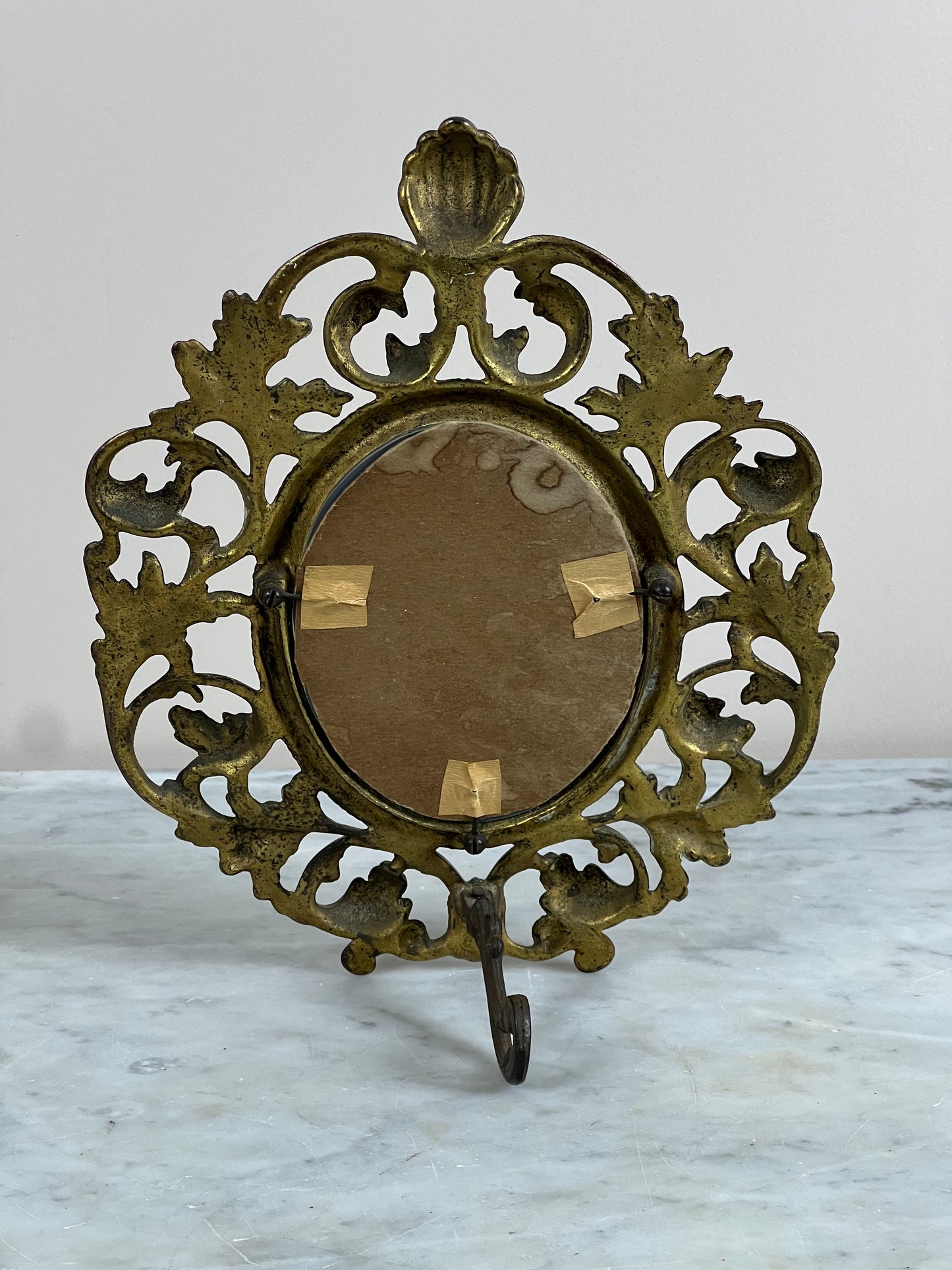 The Antique Victorian Brass Acanthus Leaf Table Mirror by Pearl City Antiques features ornate leafy scrollwork, an upright oval center with brown cardboard held by tape, and a decorative shell motif on its vintage brass frame.