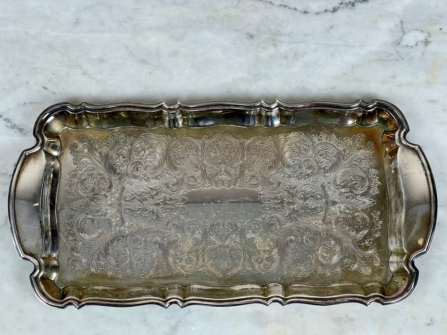 The Antique Silver Plate Serving Tray by Pearl City Antiques showcases ornate engraved floral and scroll patterns, scalloped edges, and side handles, elegantly displayed on a light marble surface.