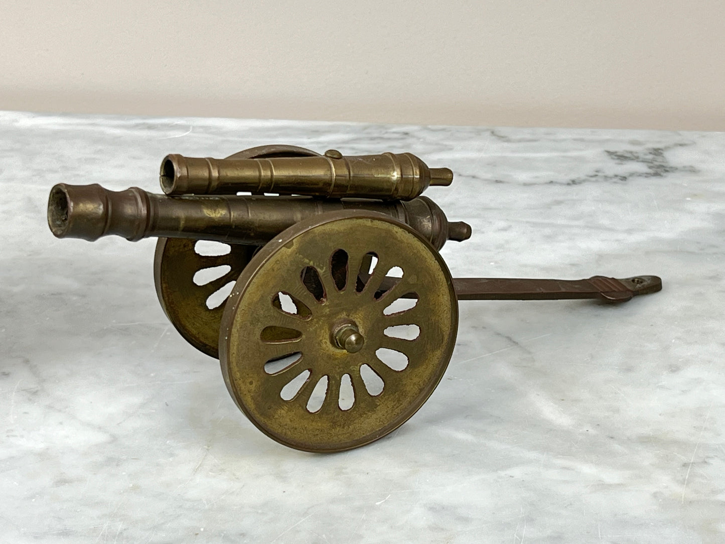 The Pair of Cast Brass Double Cannon Figurines by Pearl City Antiques feature large spoked wheels and long handles, creating striking military decor when displayed on a marble surface against a plain background.