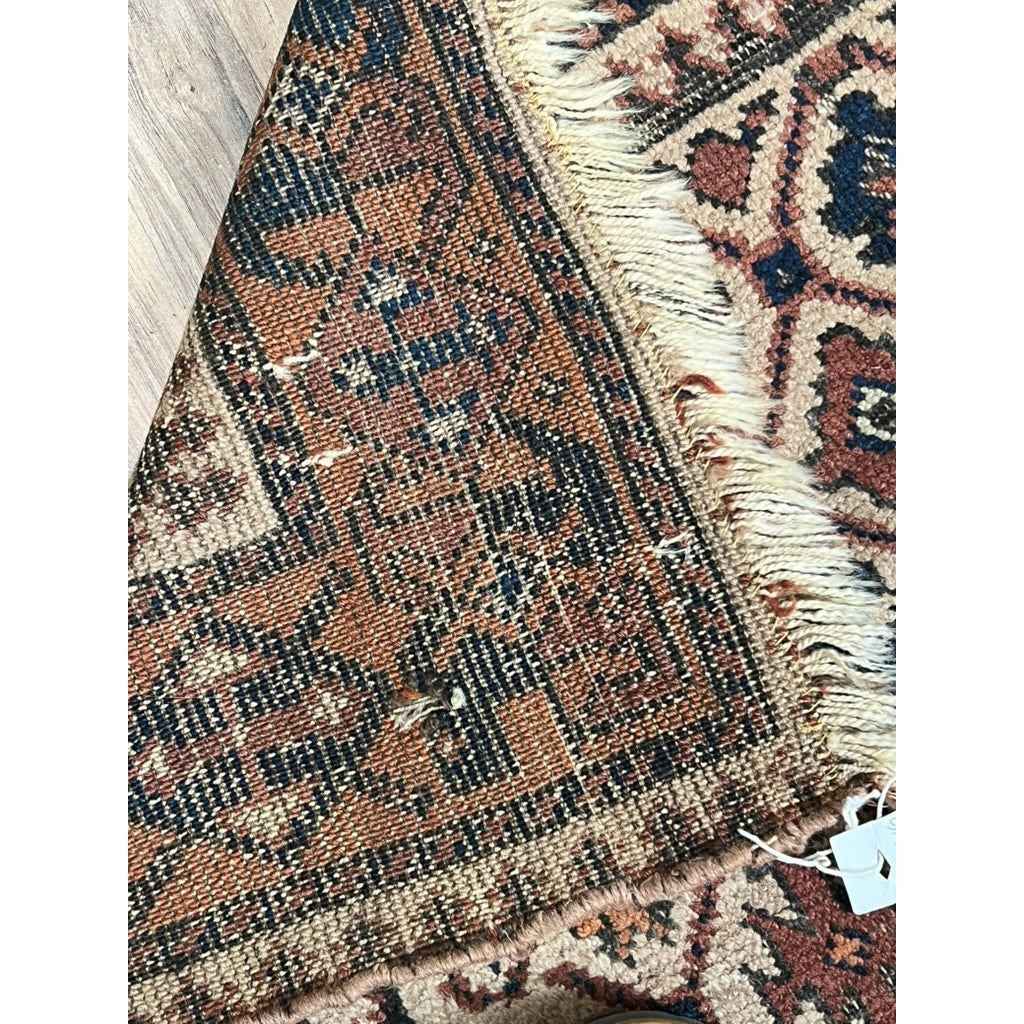 A detailed view of an Antique Baluch Tribal Rug with Caucasian influence (Northeast Persia/NW Afghanistan, mid-19th c., 3’ x 4’2”) shows geometric patterns in brown, black, and cream wool with frayed edges and a small white tag on a wooden floor.
