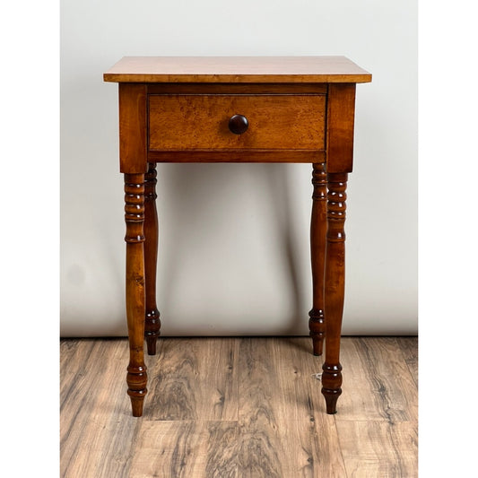 An Antique American Sheraton Birdseye Maple One Drawer Stand from the mid-19th century by Pearl City Antiques is presented here, featuring a single drawer with a round knob, turned legs, and a polished finish that exemplifies classic craftsmanship—this elegant piece stands on a wood floor against a plain light background.