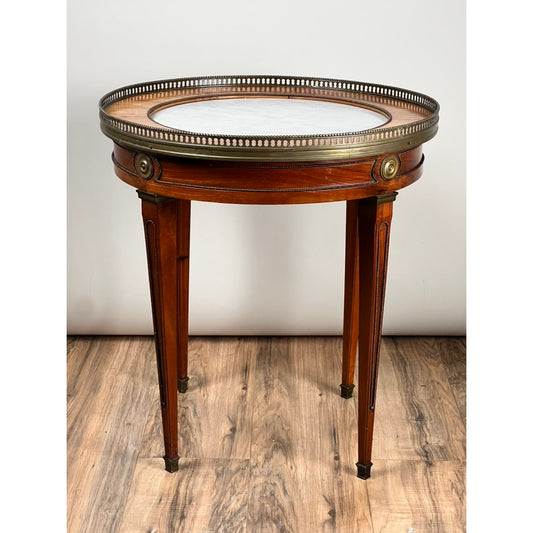 The Antique French Louis XVI-Style Mahogany Marble Top Bouillotte Table by Pearl City Antiques, dating from the early 20th century, features a round mahogany base with tapered legs, a white marble top, and elegant brass gallery edging. Reminiscent of neoclassical French design, this accent table stands gracefully on a wood floor against a plain white background.