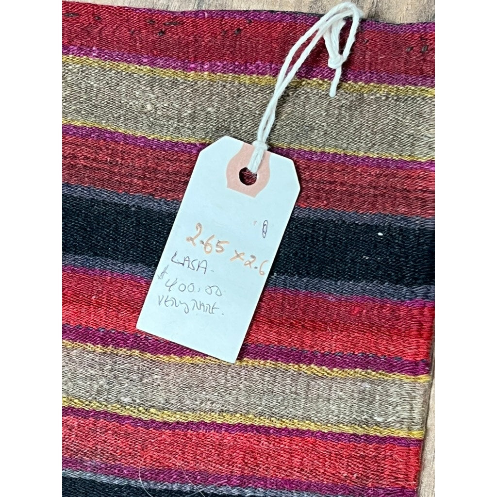 A close-up of an antique Tibetan/Nepali tribal flatweave rug (late 19th c., 2’6 x 2’7) shows red, black, pink, and beige horizontal stripes with a handwritten white tag displaying dimensions and the word "Lasa." Brand unknown.