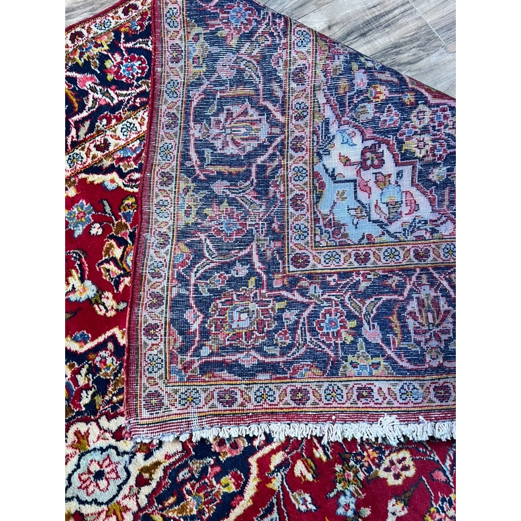 A close-up of a Vintage Hand-Knotted Persian Kashan Wool Gallery Rug (c. 1950–1970, 4’5 x 8’8), one corner flipped to reveal its intricate floral pattern in red, blue, white, and yellow on both sides. Brand: None/Unknown.