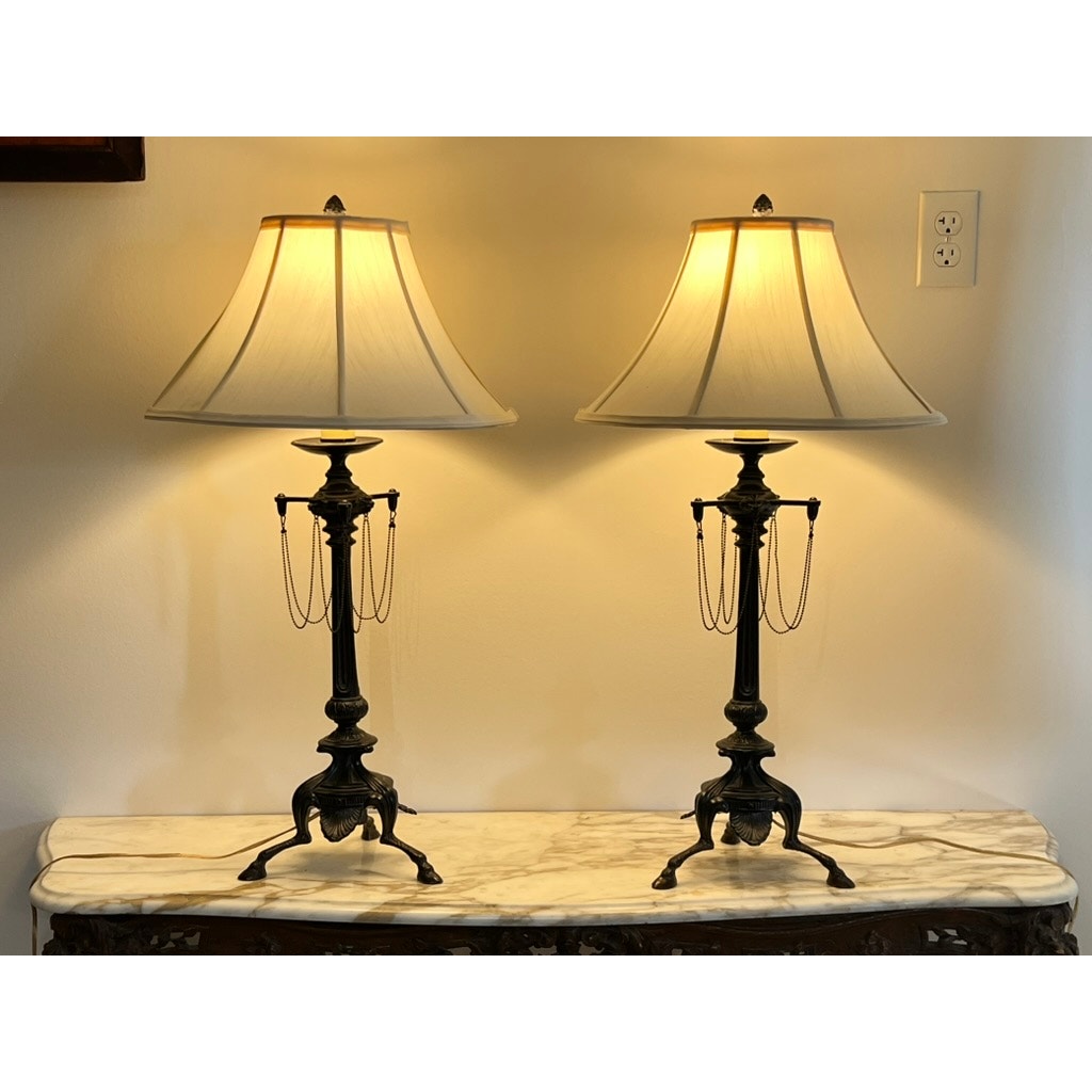 A pair of vintage French Empire Revival table lamps with oil rubbed bronze finish are illuminated on a marble-topped table against a beige wall near an electrical outlet.
