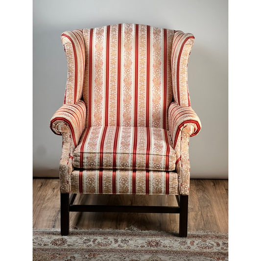 The Vintage Chippendale Style Wingback Chair by Ethan Allen from Pearl City Antiques, featuring elegant red and gold striped upholstery, wooden legs, and placed atop a patterned rug against a light-colored wall and wooden floor, brings timeless traditional charm to any space.