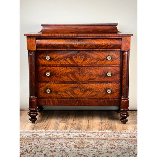 The Antique American Empire Albany Four Drawer Chest c. 1840 by Pearl City Antiques is expertly crafted from rich flame mahogany and showcases four drawers adorned with ornate gold knobs, carved side columns, and turned feet. Set against a white wall on a wood floor with a patterned rug in front, this timeless 1840s furniture piece exudes classic elegance.