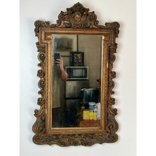 The Antique Italian Regency Style Wall Mirror with Cherub Crest by Pearl City Antiques, featuring ornate giltwood and gold detailing, hangs on a wall and reflects a person taking a photo along with a microwave, kitchen items, and shelves in the background.