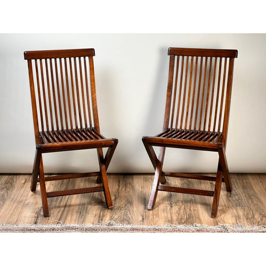 A Pair of Vintage Mid-Century Modern Style Teak Folding Chairs by Pearl City Antiques, featuring vertical slats on the backrests and seats, are displayed side by side on a wooden floor against a plain light-colored wall. Their clean lines and understated elegance evoke a touch of Scandinavian design.
