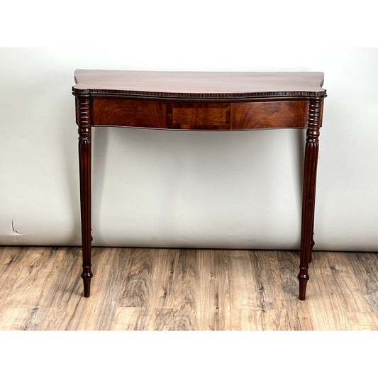An Antique Mahogany Sheraton Game Table with a serpentine front, c. early 19th century, stands on a wood floor against a plain light-colored wall.