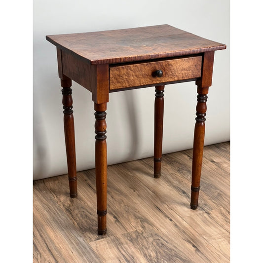 The Antique Sheraton One Drawer Tiger Maple Stand by Pearl City Antiques, dating to the mid-19th century, features beautiful tiger maple construction, four turned legs, and a single drawer with a round knob. It is shown standing on a wood floor against a plain wall.