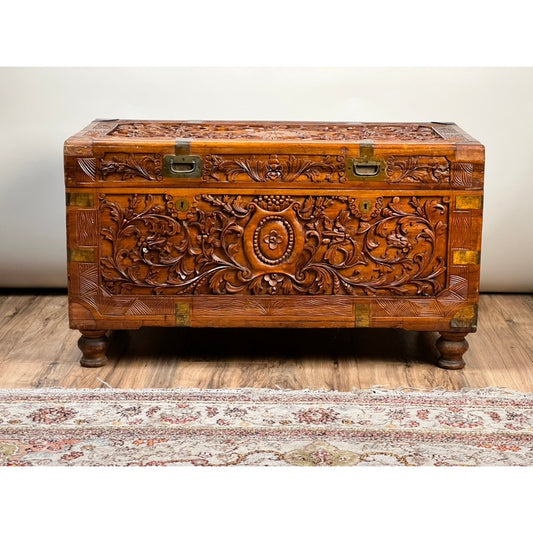 The Chinese Export Carved Camphor Wood Dowry Chest by Pearl City Antiques features intricate floral and geometric carvings, brass hardware, and short, rounded legs, elegantly displayed on a wooden floor beside a patterned rug.