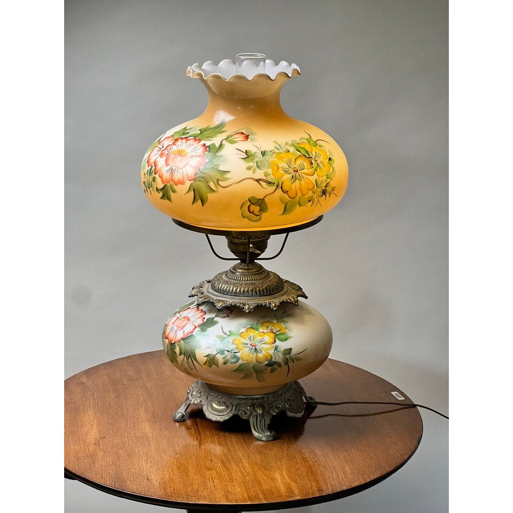 The Vintage Hand-Painted Floral Milk Glass Hurricane Parlor Lamp sits on a round wooden table against a plain gray background, featuring floral designs on its milk glass and ornate metal accents, with a glowing upper shade.