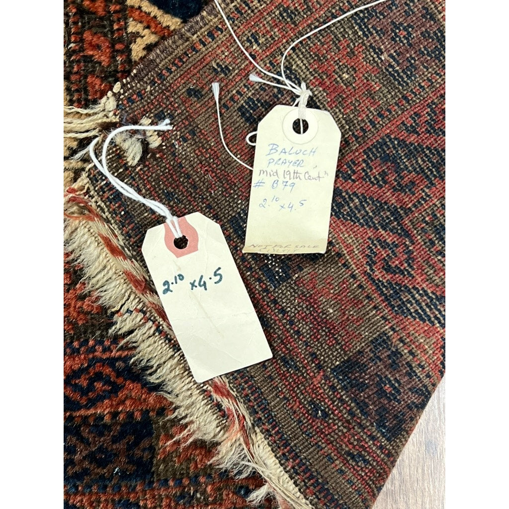 Two handwritten tags rest on a patterned rug: one shows the size, 2’10” x 4’5”, and the other notes “antique Baluch prayer rug.” The visible fringe and weaving belong to an Antique Baluch Prayer Rug, NE Persia, mid-19th c., 2’10” x 4’5”.