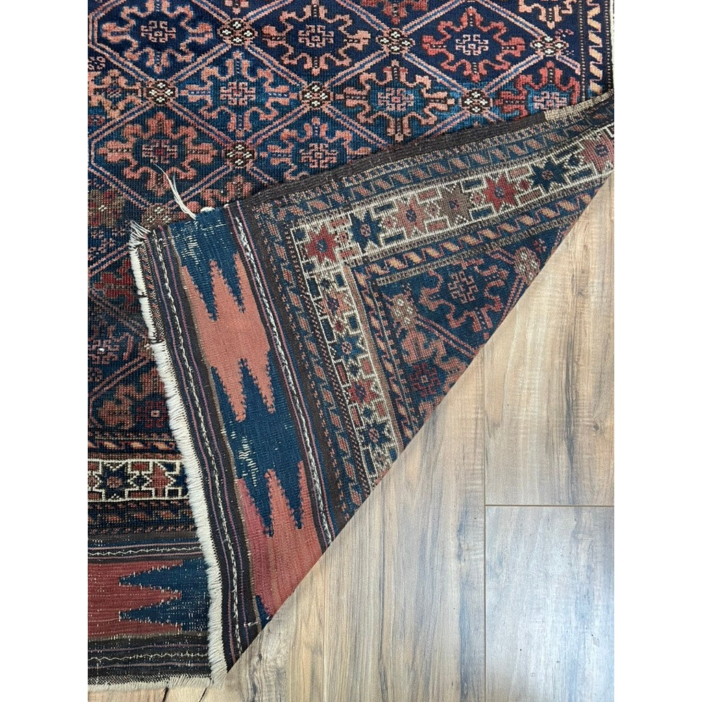 An Antique Tribal Baluch Rug with a diagonal lattice field (Northeast Persia, late 19th century, 3’1 x 5’2) in dark red, navy blue, and beige lies partly folded on a wooden floor, showing its intricate underside and fringed edge.
