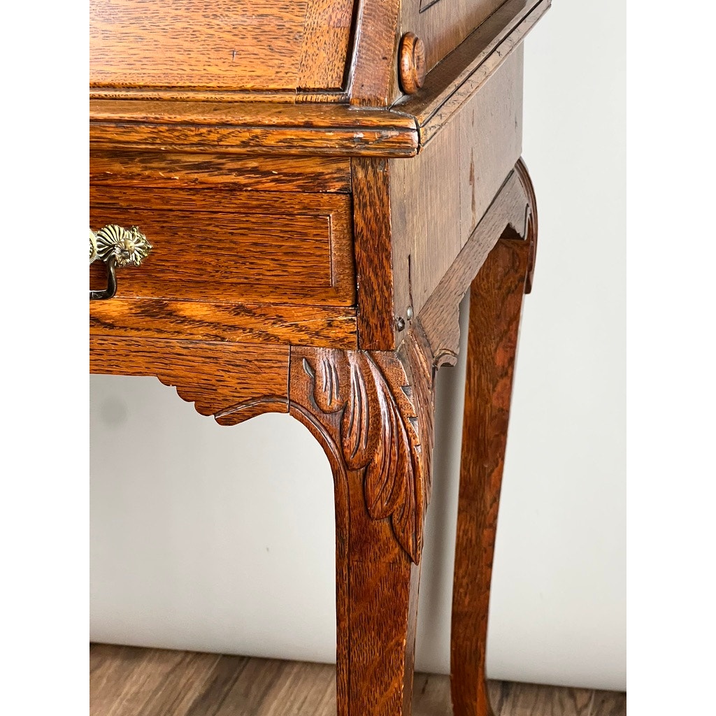 Close-up of an Antique Victorian Oak Ladies Writing Desk c. 1900, featuring ornate leg carvings, a brass handle, and a polished oak finish, set on a wood floor against a plain light background.