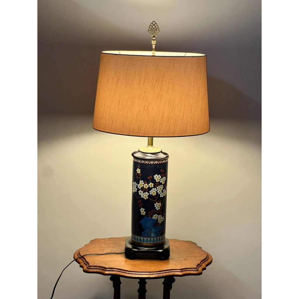The Vintage Japanese Cloisonné Cylinder Table Lamp by Knob Creek, featuring a beige shade and floral base, sits on a small wooden table against a plain wall. The lamp is turned on, casting a warm glow.
