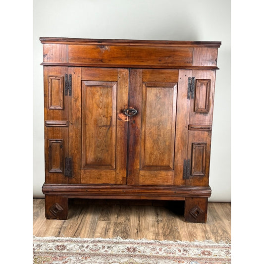 An Antique Continental Pine Cupboard, probably German from the early 18th century by Pearl City Antiques, featuring two paneled doors with metal hinges and a latch, stands on square legs atop a wooden floor, with part of a patterned rug visible below.