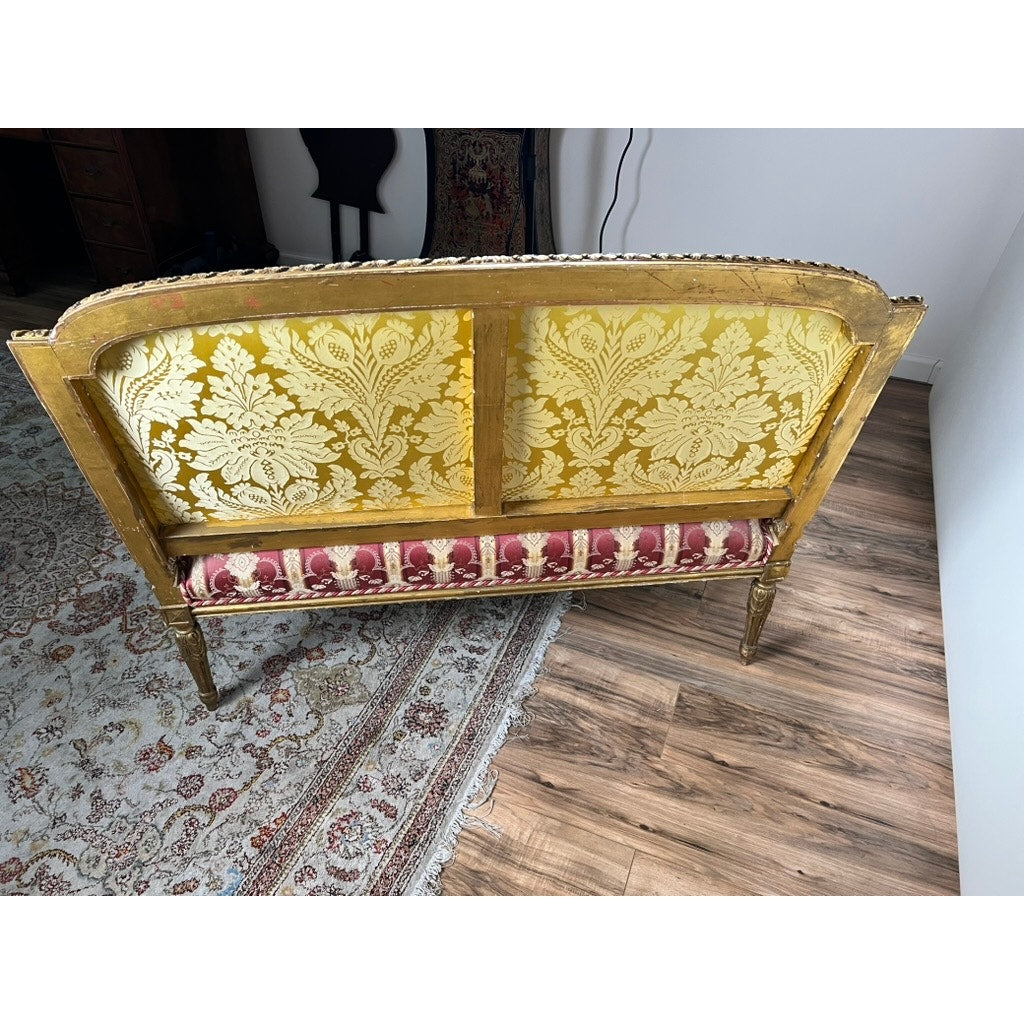 An Antique Louis XVI Style Giltwood Settee in silk damask (late 19th century) sits on a patterned rug over a wooden floor near a white wall, featuring gold floral upholstery on the backrest and a red-and-gold seat.