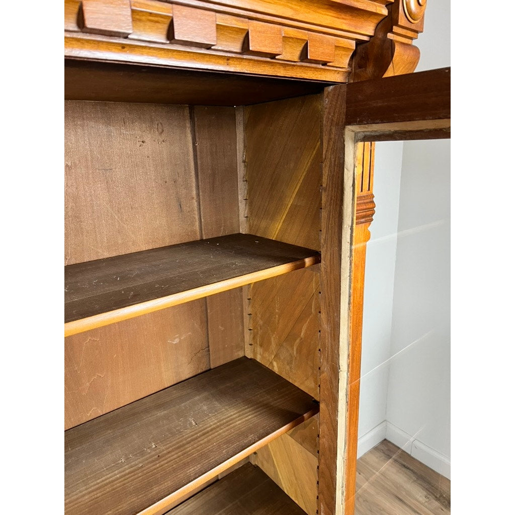 Close-up of an Antique Eastlake Victorian Walnut Triple Bookcase c. 1880 with empty shelves, decorative top trim, and a glass door panel. This striking piece stands on a wooden floor against a white wall.