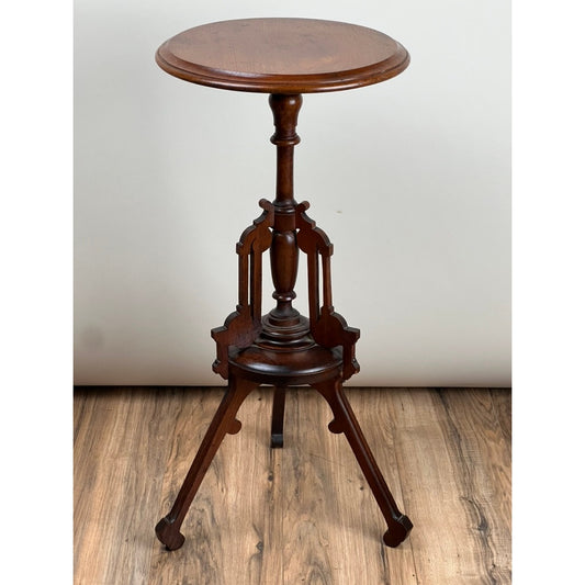 An Antique Victorian Walnut Candle Stand by Pearl City Antiques (c. Late 19th Century), featuring a round wooden top, an ornate central column, and three gracefully curved legs, stands on a wood floor against a plain light-colored wall.