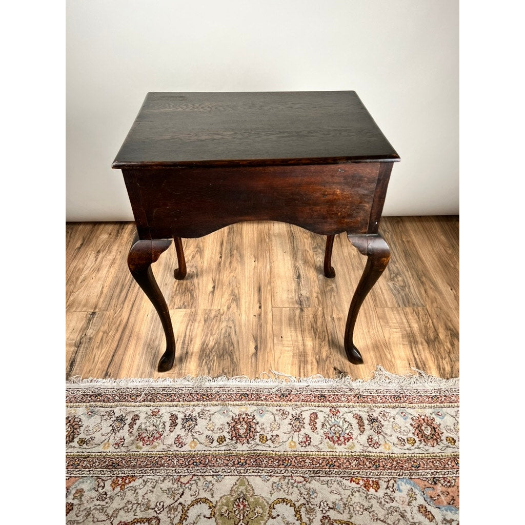 An Antique Queen Anne Style George III Oak One Drawer Stand from the late 18th century stands on a wood floor beside a patterned rug, set against a plain, light-colored wall. Brand: None/Unknown.