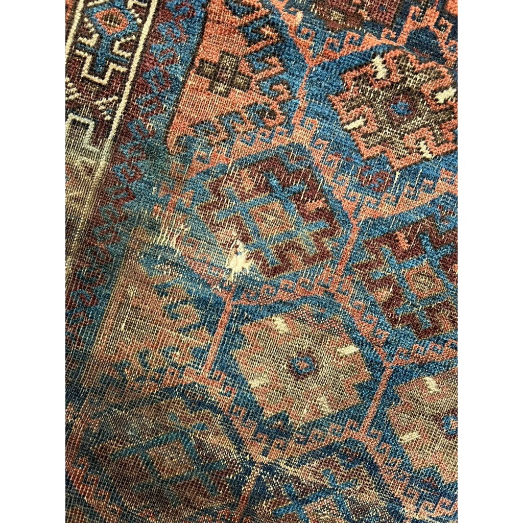 A close-up of an Antique Baluch Prayer Rug from the Khorosan region, NE Persia (early 20th c., 3’ x 5’3), shows geometric blue, rust, and beige patterns and a frayed area exposing the underlying material.