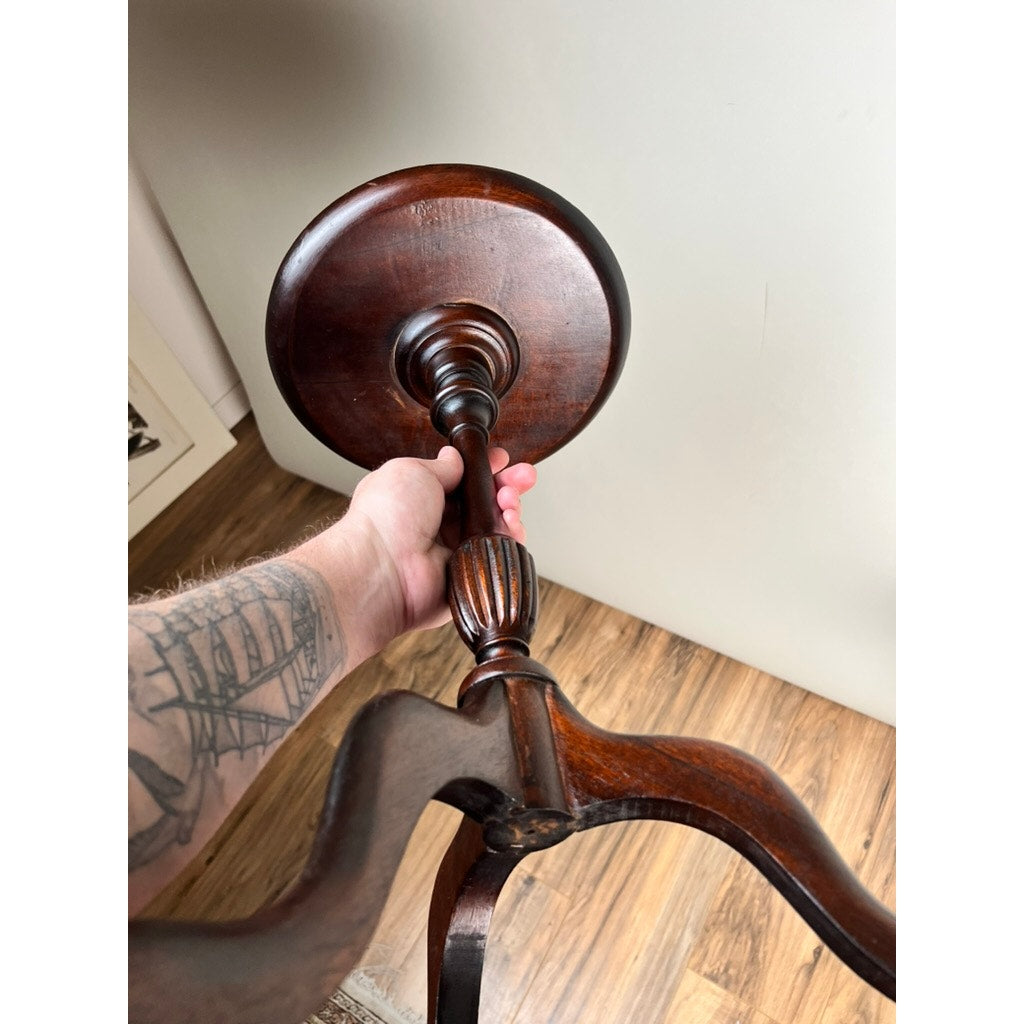A tattooed arm holds up a Vintage Match Pair Diminutive Walnut Candle Stand c. 1940 upside down, displaying its pedestal base and three curved legs, set against a wooden floor and light-colored wall—ideal for vintage decor lovers.