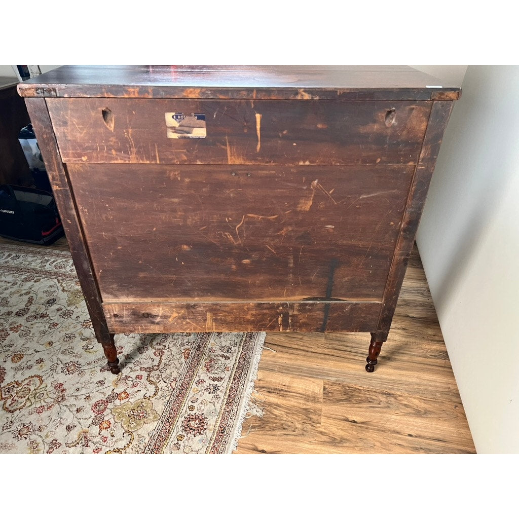 An Antique George III Sheraton Flame Mahogany Chest of Drawers (early 19th century) with small wheels stands against a white wall on a patterned rug and wood floor.