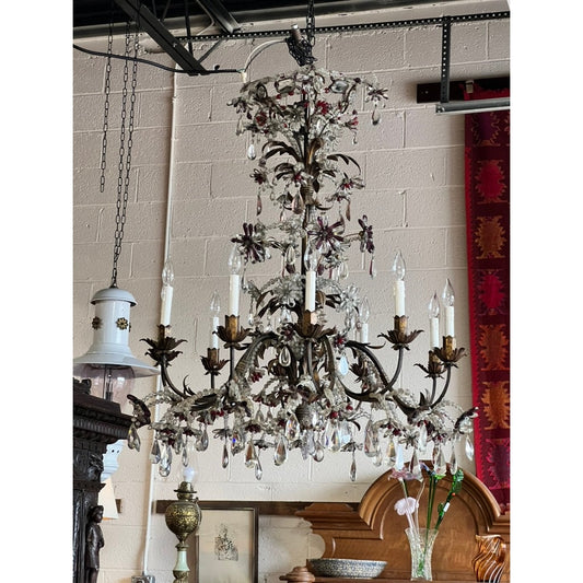 The Antique Late 19th Century French Rococo Tole Amethyst and Crystal Ten Light Chandelier by Pearl City Antiques, adorned with sparkling prisms and floral accents, hangs from the ceiling amidst antique furniture and decor set against a light brick wall.