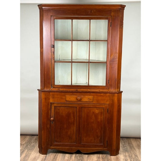 The Antique Pennsylvania Nine Panel Glass Door Corner Cupboard c. Early 19th Century by Pearl City Antiques is a wooden cabinet featuring a distinctive nine panel glass door on the upper section, a single drawer in the middle, and two solid doors below. It stands gracefully on a wood floor against a plain background.