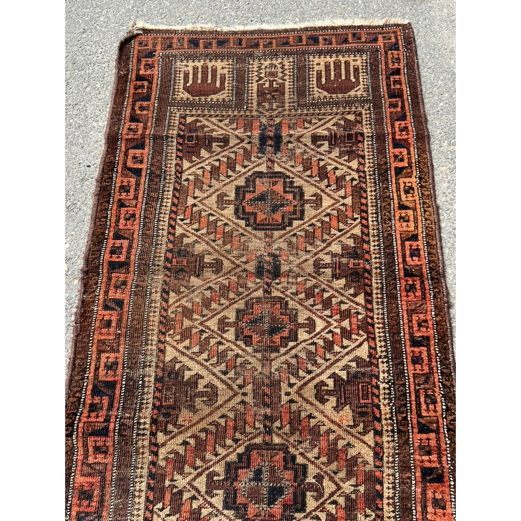 An Antique Northeast Persian Baluch Prayer Rug (c. mid-19th century, 2’5 x 4’6) with geometric and diamond motifs in brown, tan, rust, and black displays intricate borders—a striking tribal textile shown on concrete. Brand: Unknown.