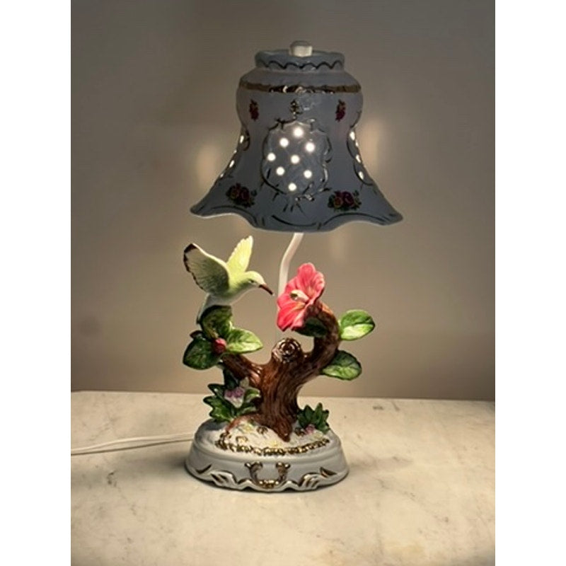 The Vintage Lena Liu Porcelain Hummingbird Accent Lamp features a tree branch base, a colorful hummingbird sipping from a pink flower, and a floral-patterned shade. It glows warmly, adding floral charm to any light-colored surface.