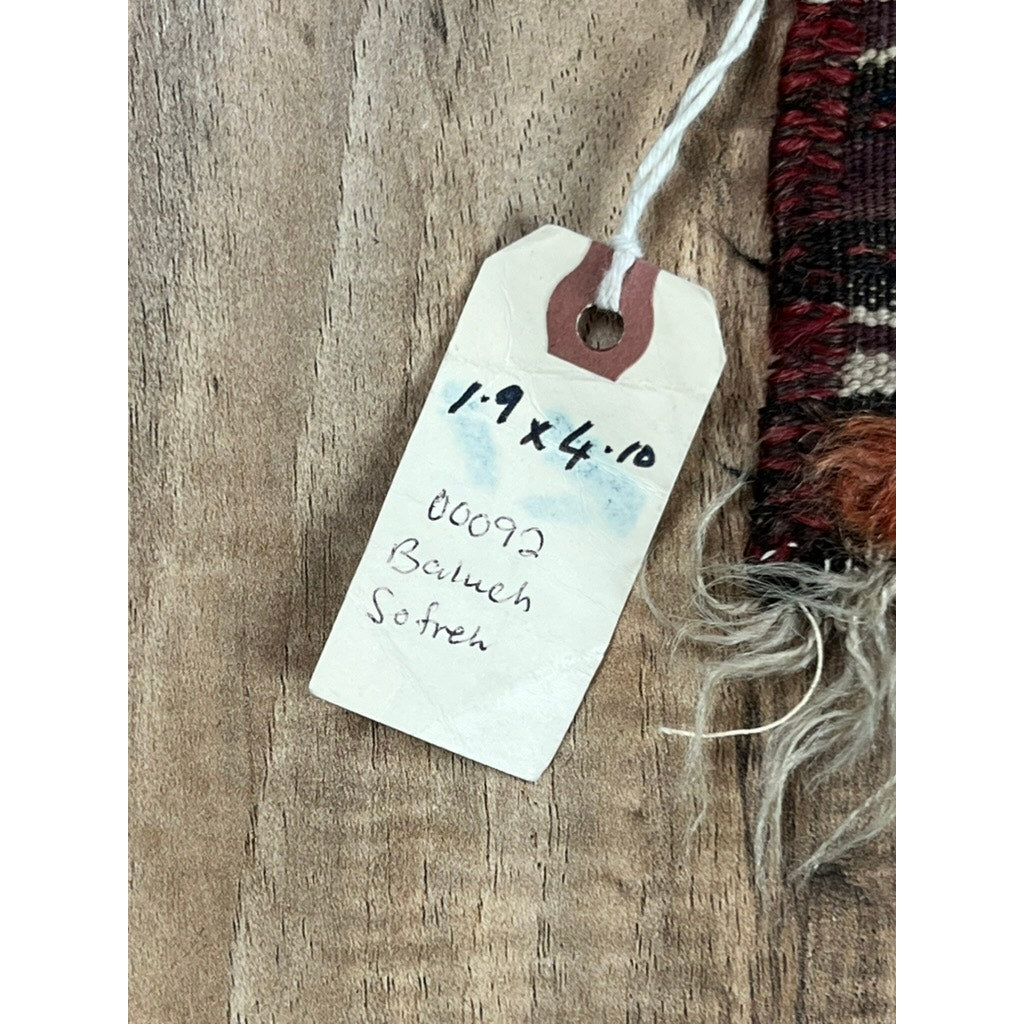 A close-up of a handwritten tag on woven fabric reads “1’9 x 4’10, 00092, Baluch Sofreh,” identifying an Antique Baluch Sofreh Flat Weave Rug c. Late 19th Century, shown on a wooden surface.