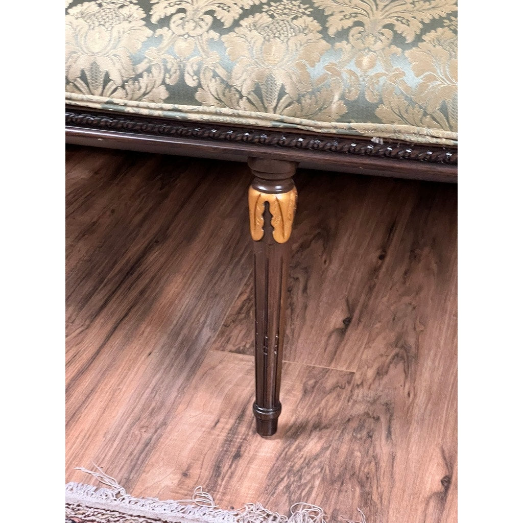 Close-up of a Vintage Louis XVI Style Settee in green damask upholstery with a floral pattern and ornate wooden leg with gold accents, set on a wooden floor near a fringe rug. Brand: None or Unknown.