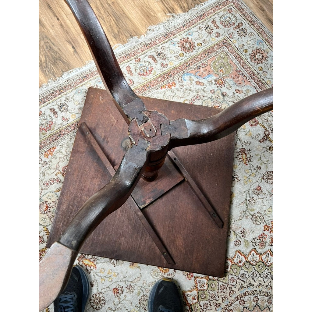 An Antique Queen Anne Mahogany Tilt Top Breakfast Table (late 18th c., maker unknown) lies upside down on an ornate rug, with its central leg and three curved supports damaged and detached; sneakers are partly visible at the bottom edge.