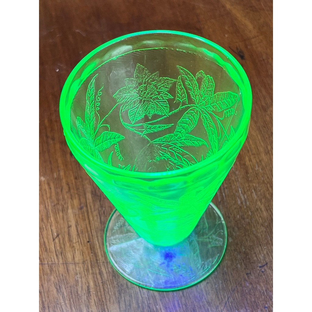 An Antique Jeannette Glass Co. Green Uranium Floral Poinsettia 5-Piece Beverage Set from the 1930s features glowing glassware under UV light, beautifully displaying intricate engravings on a wooden surface.