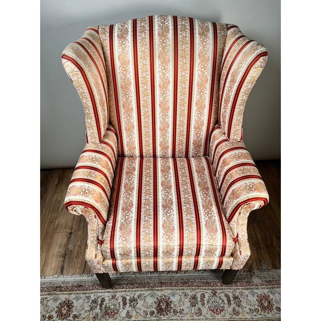 The Vintage Chippendale Style Wingback Chair by Ethan Allen features ornate wooden legs and traditional red, beige, and gold floral-striped upholstery, positioned on a hardwood floor beside a decorative rug.
