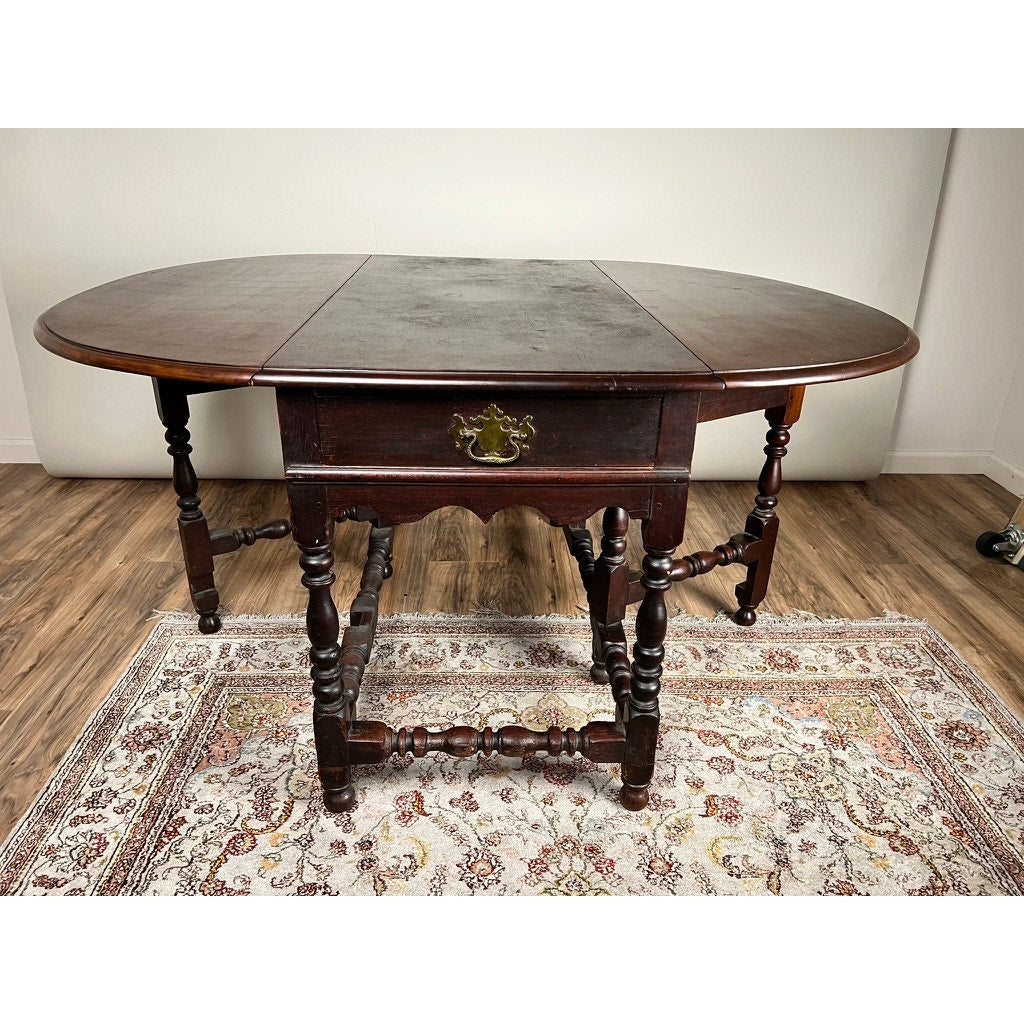 An Antique English William & Mary Walnut One Drawer Gateleg Dining Table, c. early 18th century, stands on a patterned rug in a room with light walls and wood floors.