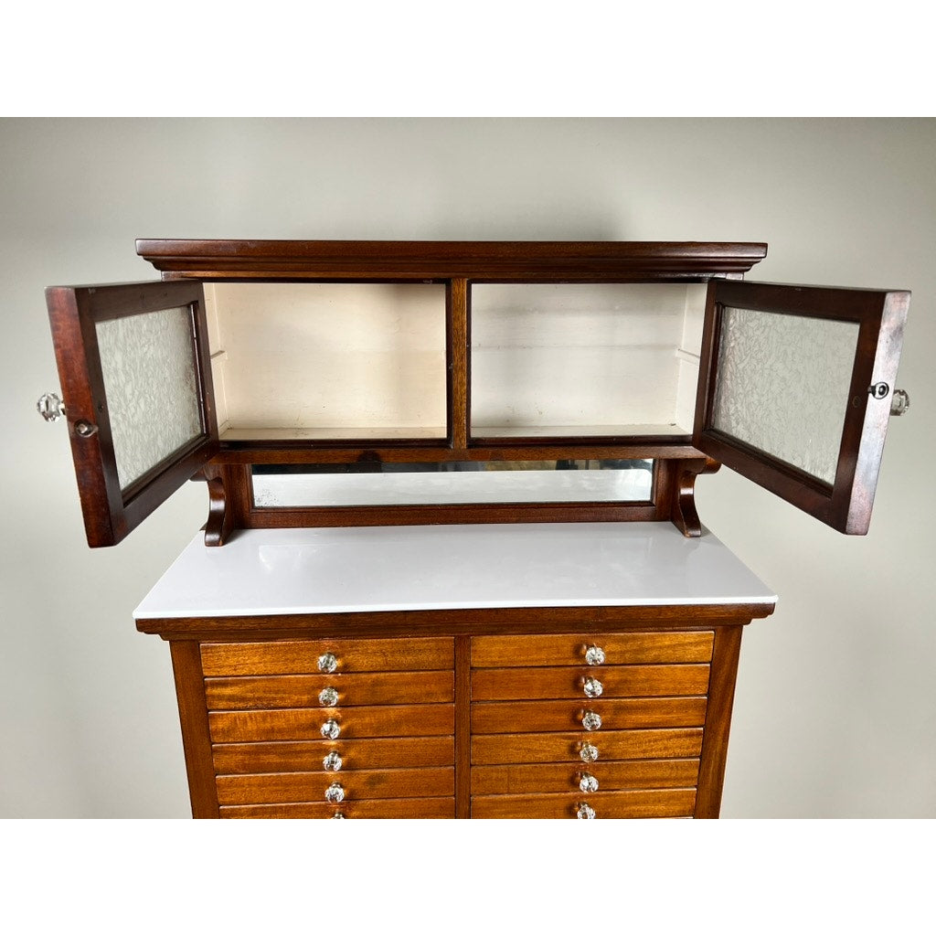 An Antique Mahogany Multi-drawer Dental Cabinet by The American Cabinet Co. (early 20th c.) features two glass-front upper doors, empty shelves, a white countertop, and drawers with round metal knobs, set against a plain light-colored wall.