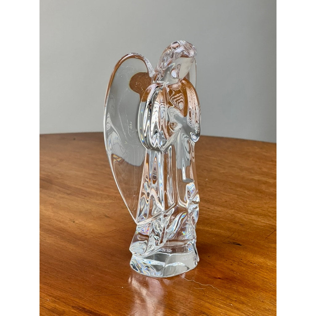 The Vintage Baccarat Crystal Angel by Baccarat stands with wings and folded hands on a polished wooden surface against a plain gray background.