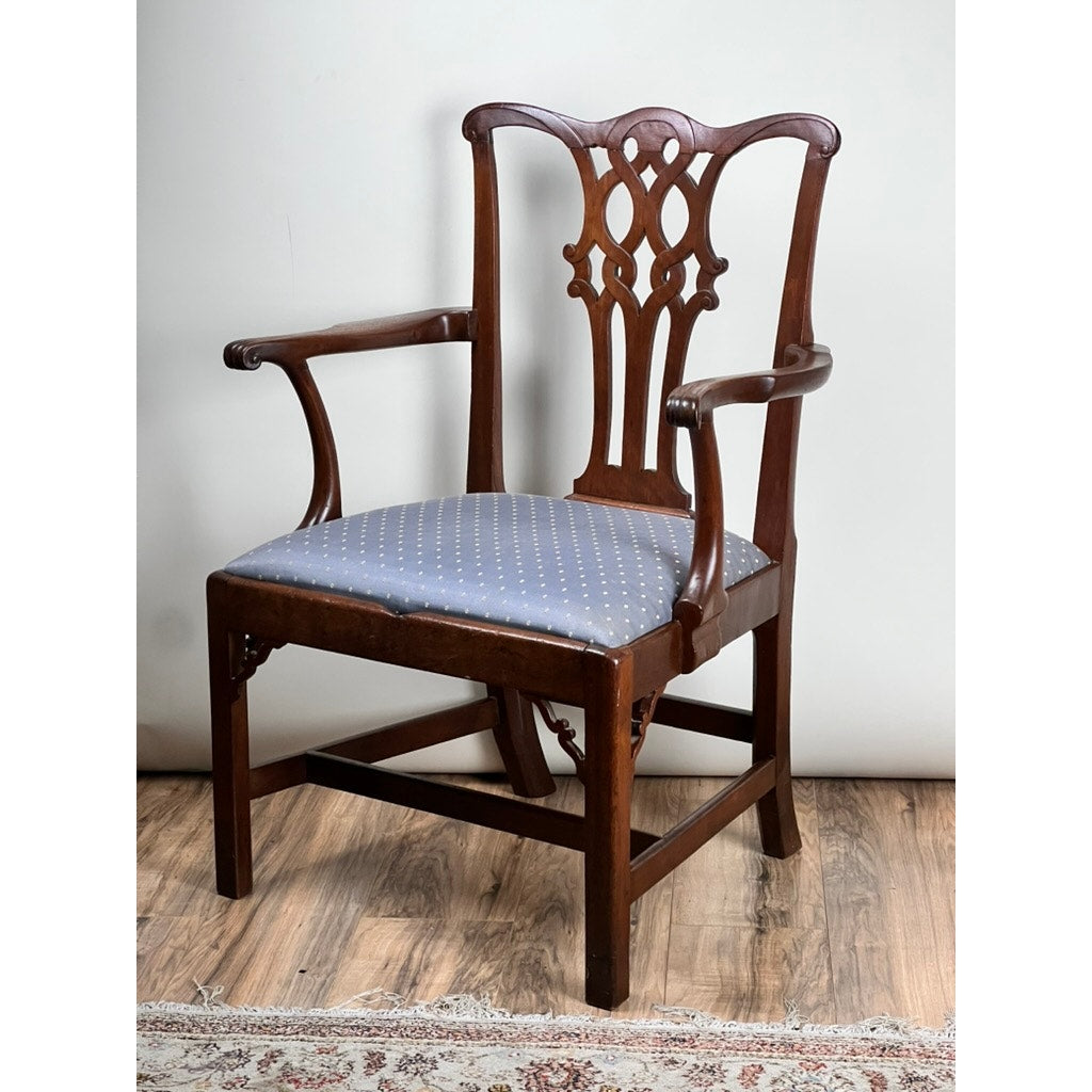 An Antique Chippendale Walnut Open Arm Chair c. Late 18th Century, featuring a carved back and blue dot upholstery, is placed on a wooden floor beside a patterned rug and in front of a plain wall.