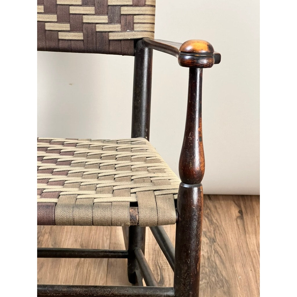 Close-up of an Antique Shaker #6 Tape Seat Rocker by Mt. Lebanon Shakers (late 19th century), showing a woven beige and brown seat and backrest, one armrest, and part of the leg on a wood floor against a light wall.