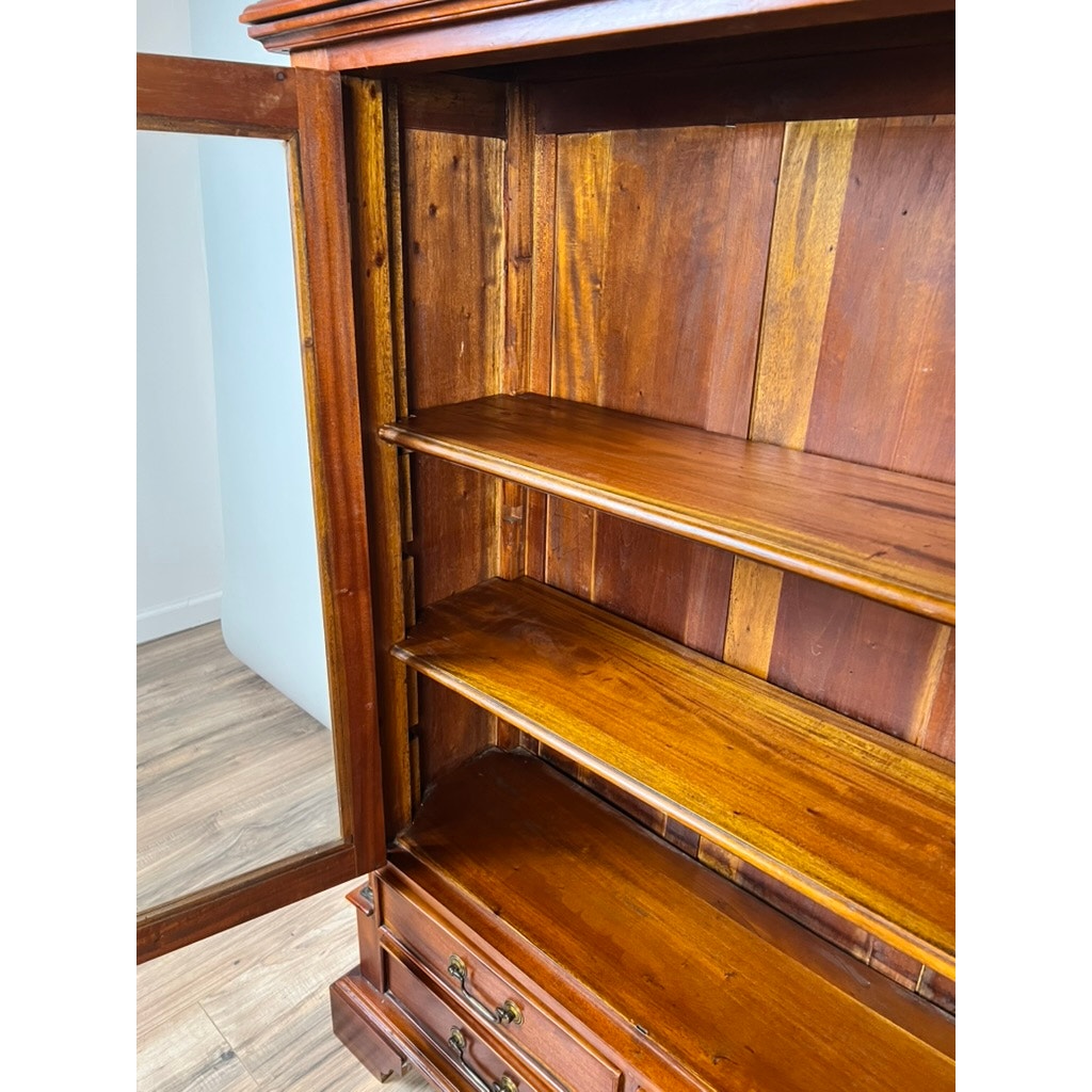 The Vintage Victorian Style Glass Door Mahogany Bookcase stands empty, featuring a mirrored door, open shelves, and two bottom drawers. It is set on a light wood floor beside a white wall.