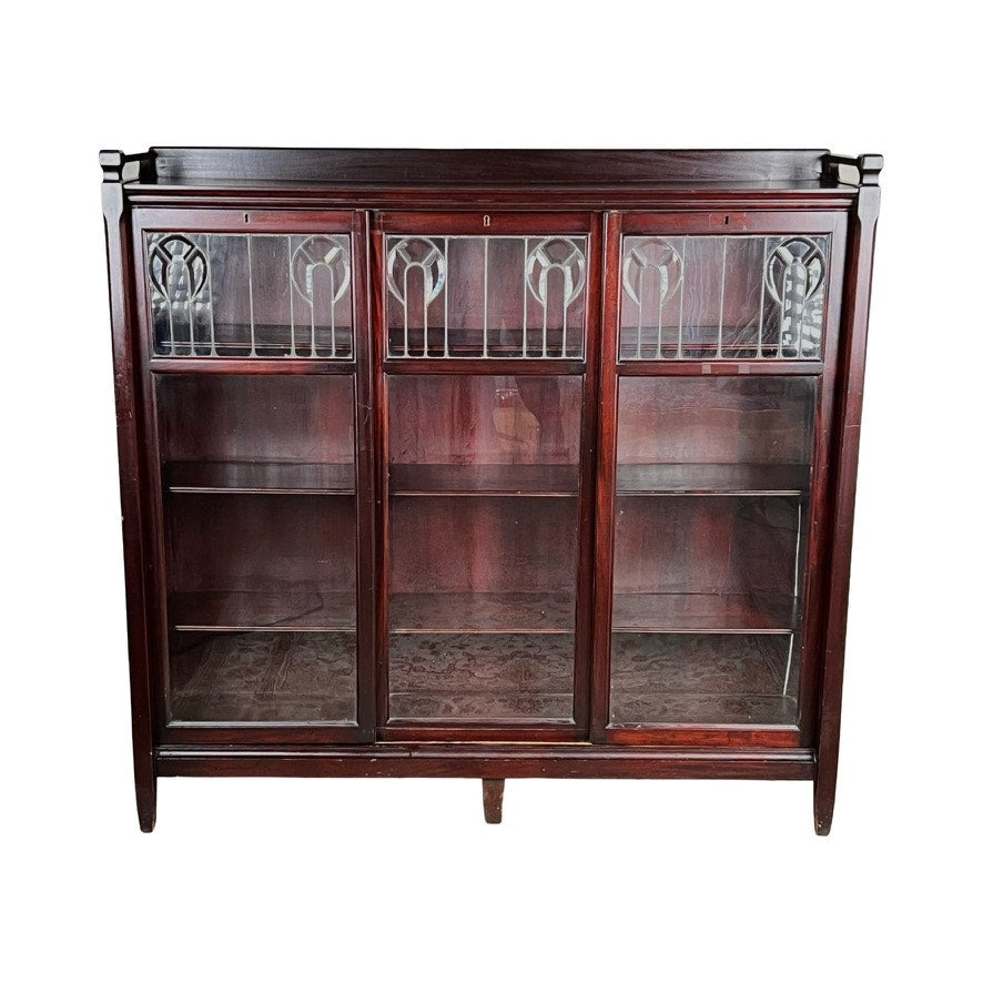 The Antique Arts & Crafts Leaded Glass Mahogany Bookcase c. 1920 features dark mahogany, three glass doors with decorative leaded glass, and interior shelves, giving it a vintage antique look. Brand: None/Unknown.
