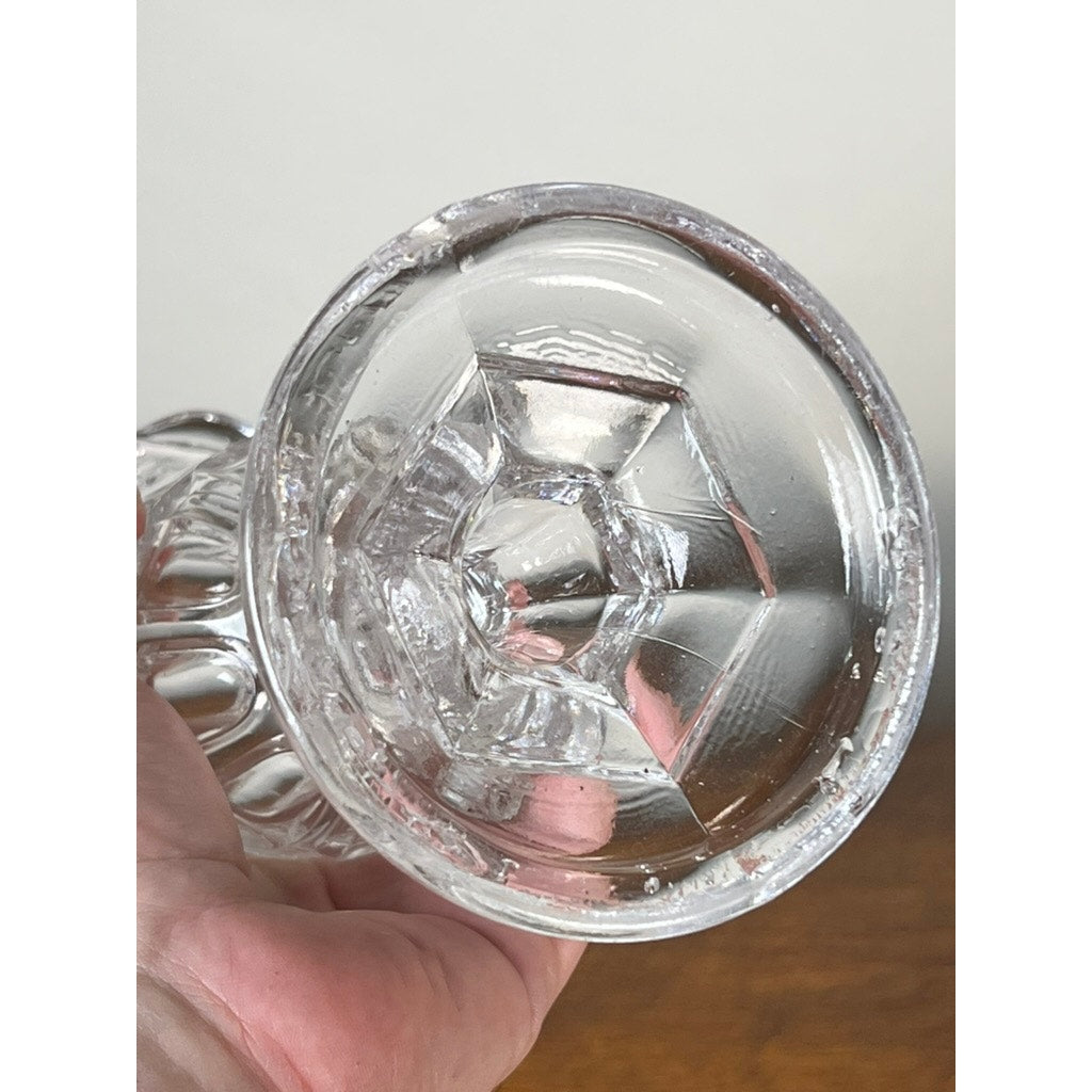 A hand holds an Antique Boston & Sandwich Glass Co. Gothic Arch Pressed Glass Celery Vase (c. 1860–1880) with a hexagonal pattern and textured rim, viewed from the bottom. The background is plain and out of focus. Brand: Sandwich Glass.