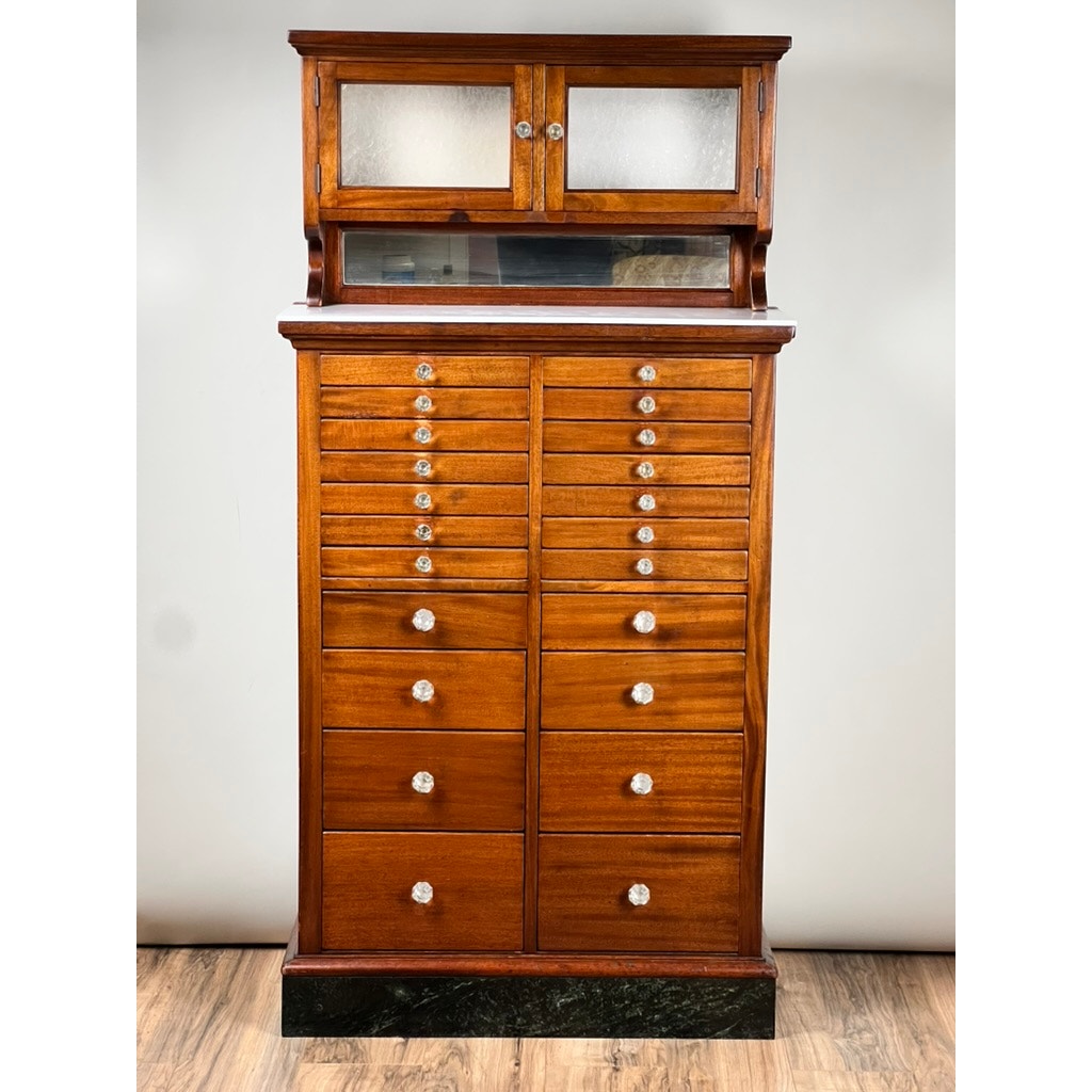 An Antique Mahogany Multi-drawer Dental Cabinet by The American Cabinet Co., early 20th century, featuring a glass-fronted top, shallow display shelf, and dual columns of round-knobbed drawers; shown on a black base against a plain background.