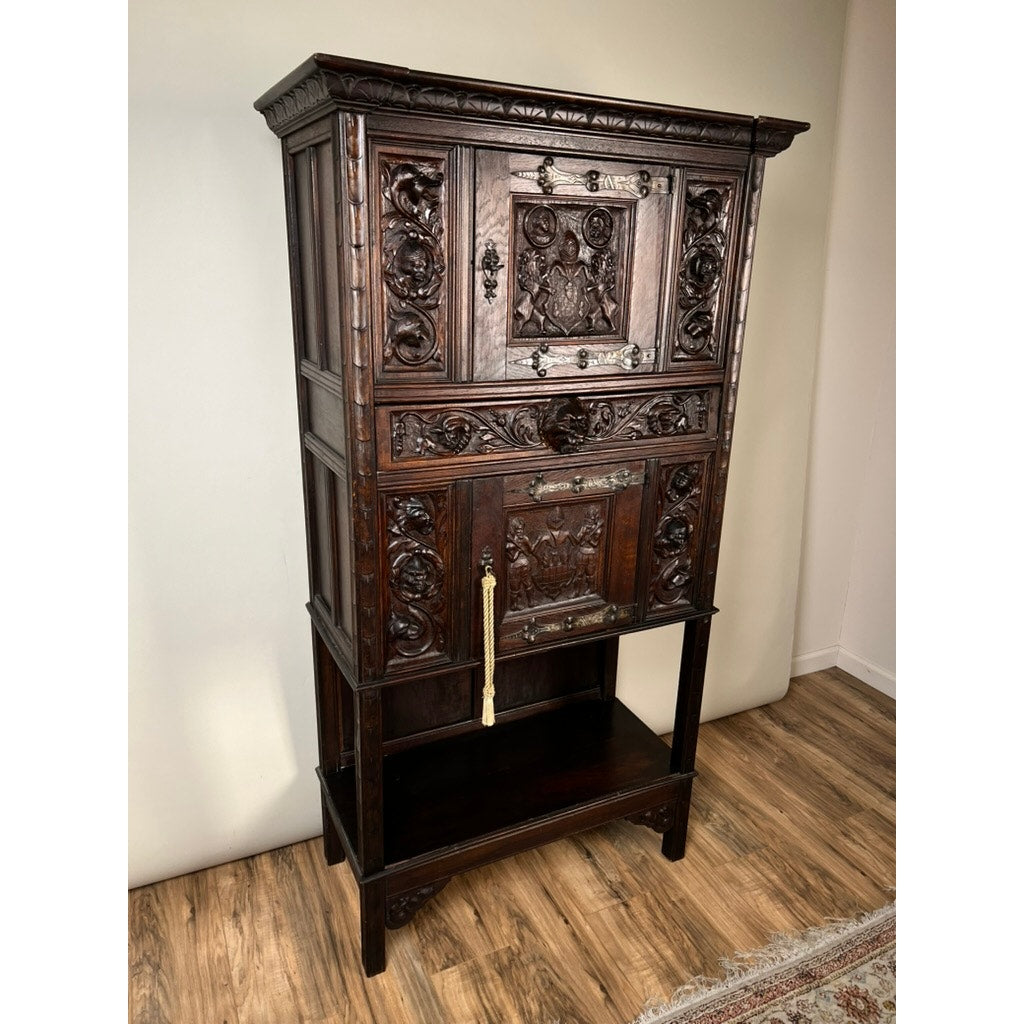 An antique 19th-century French Breton-style carved oak cabinet featuring deeply carved knights, ornate panels, a brass handle, four legs with a lower open shelf, stands on a wooden floor near a light-colored wall.